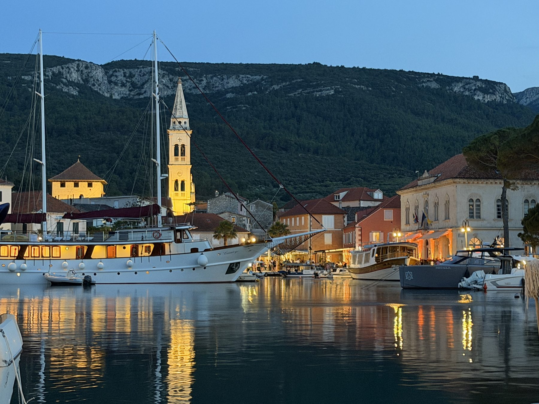 A Dalmatian harbor at blue hour, its bell tower and moored boats lit and reflecting on the water